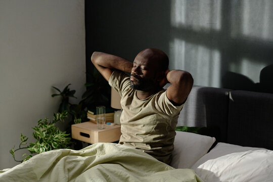 High angle shot of African American man sitting in bed stretching his muscles after sleep
