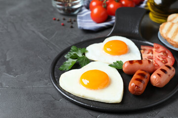 Delicious breakfast with heart shaped fried eggs and sausages on dark grey table