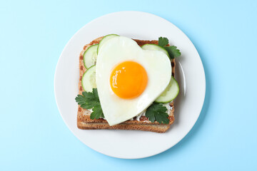 Plate of tasty sandwich with heart shaped fried egg on light blue background, top view