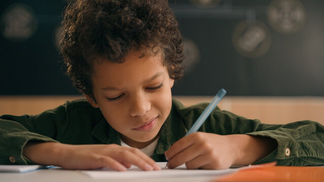 Little African American guy boy writing notes task class lesson primary school write in copybook kid schoolboy son child pupil student learning listen teacher studying education sitting at table desk