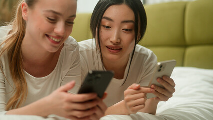 Two happy smiling multiracial friends multiethnic women diversity girls female Caucasian Asian Japanese Chinese relaxing on bed together using mobile phones discuss news online social media talking