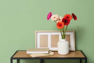Beautiful gerberas in vase, books and photo frame on table near green wall in room