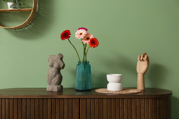 Beautiful gerberas in vase and statuettes on cabinet near green wall in room