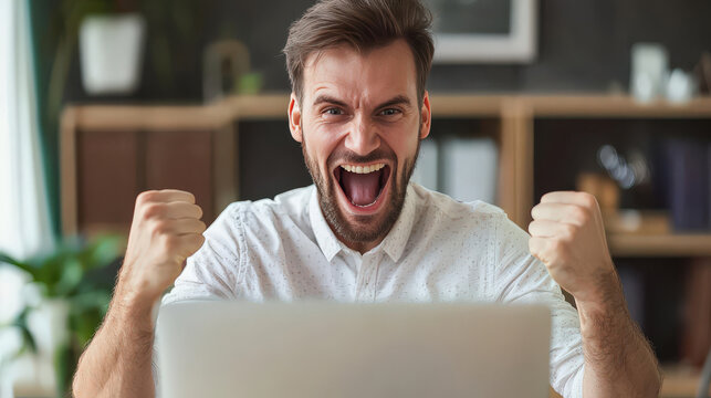 Excited Young Man Celebrating a Victory with Fists Raised in Front of a Laptop in a Modern Workspace Filled with Plants and Natural Light