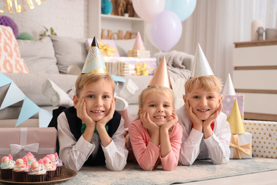 Cute little happy kids in party hats with cupcakes and gift boxes celebrating birthday at home - Powered by Adobe