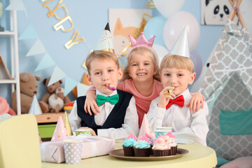 Little happy kids with cupcakes and gift box blowing whistles while celebrating birthday at table in children's room