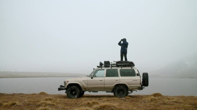 Young Man climbs onto the roof of car and looks into the distance. SUV is parked close to serene and foggy lake, surrounded by majestic mountain scenery