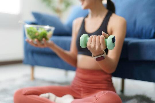Young woman lifting light dumbbell and holding bowl of fresh salad, demonstrating commitment to healthy habits and fitness - Powered by Adobe