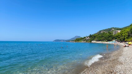 The wave, foaming, rolls onto the pebble beach of the seashore. The mountains in the bay are covered with forest. People are swimming in the sea. Pine trees and buildings stand on the shore. Sunny