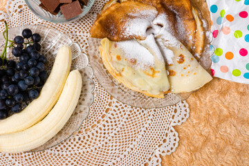 Thin pancakes dusted with powdered sugar are presented on a delicate doily alongside fresh bananas,...