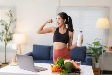 Young asian sportswoman showing muscles and holding milk bottle after online workout with laptop at home