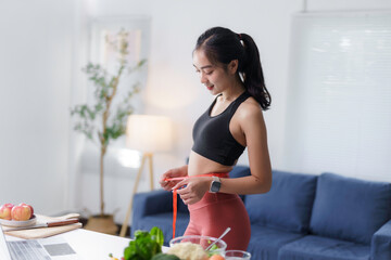 Happy asian woman measuring her waistline with tape measure while preparing healthy food in kitchen...