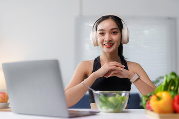Young asian nutritionist wearing headphones smiling at laptop with vegetables and fruit on table,...