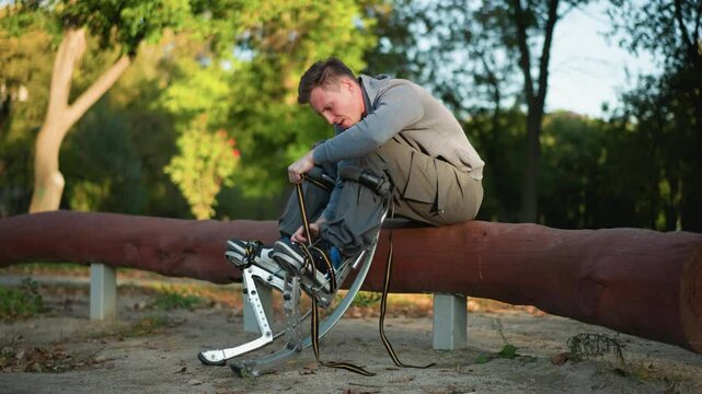 Guy sitting on log in park, wearing gray long-sleeved shirt and cargo pants, focused on adjusting spring stilt straps attached to feet, surrounded by green trees, with low camera angle