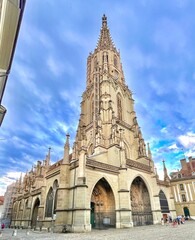 Obraz premium This image shows a tall, intricately designed Gothic-style cathedral with a prominent spire reaching towards the sky. The architecture features pointed arches, detailed stone carvings, and numerous sp