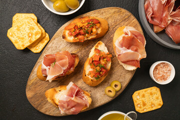 Sandwiches bruschetta with olives, bran bread, cherry tomatoes, prosciutto on wooden cutting board, dark black background surface
