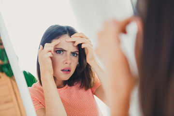 Fototapeta premium Young woman examining her skin in front of a mirror in a bright, cozy living room during the day