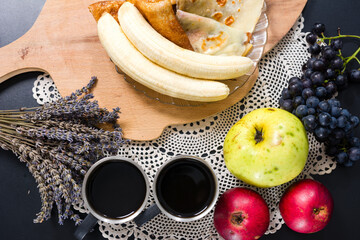 Overhead view of a cozy breakfast setting with coffee, pancakes, bananas, apples, grapes, and a sprig of dried lavender on a lace doily.