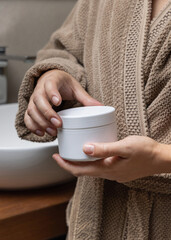 Closeup of woman in brown bathrobe holding white cream jar in hands, cosmetic mockup