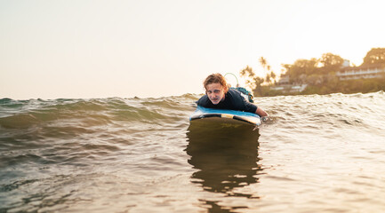 Young man paddling on surfing surfboard on surfing spot in Indian ocean. Palm grove litted sunset rays in the background. Extreme water sports and traveling to exotic countries concept.