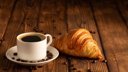A cup of fragrant black coffee and an appetizing croissant on a wooden background. Coffee beans.