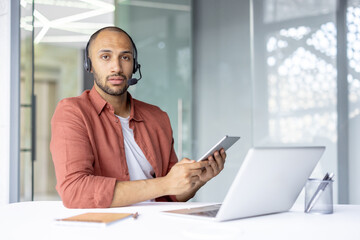 Confident customer service representative wearing a headset and using a tablet while working....