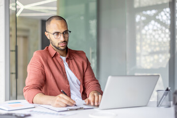 A professional sitting at a desk working with a laptop and writing notes, conveying concentration...