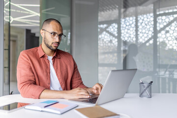 A focused individual wearing glasses works diligently on a laptop in office, conveying professionalism, productivity, and concentration, complemented by office supplies and thoughtful interior