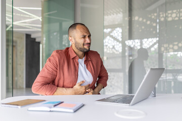 Naklejka premium A professional seated at a desk appears in discomfort while holding his stomach, expressing pain. The modern office environment emphasizes his professional context and the seriousness of situation.