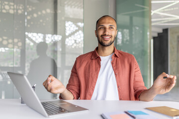 A young man in a casual shirt smiling and meditating at a desk with a laptop in a bright and modern office environment. The mood conveys calmness and balance during work hours.