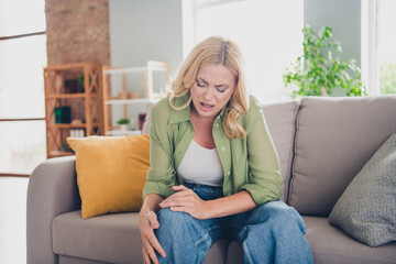 Mature woman experiencing knee discomfort while relaxing at home in a casual outfit, feeling discomfort and pain.