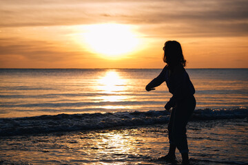 Fototapeta premium Silhouette of woman throwing stone in sea water at sunset creating golden light reflection