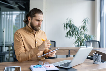 A dedicated business professional concentrates on taking notes while working on a laptop. The serene office setting adds to the productivity and professional environment.