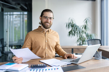 Fototapeta premium Smiling office worker sitting at desk with laptop, reviewing documents, and analyzing reports. The workspace exudes professionalism and productivity
