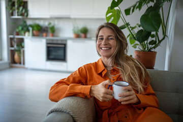 Pleased dreamy middle aged woman sits on couch relax at home holding cup coffee. Joyful female...