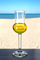 Glass of grappa on the wooden table in the beach bar. Picturesque background with sand and sea. Glassware with the drink. 3d rendering.