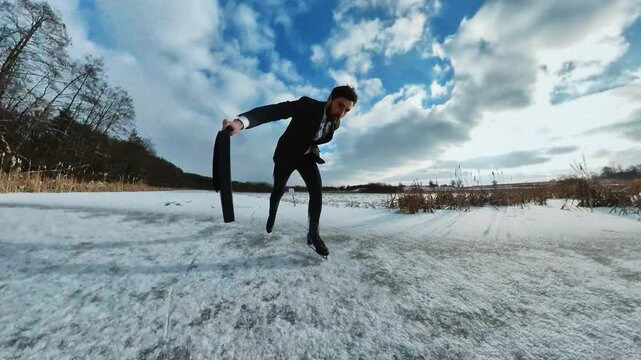 Footage of ice skating young handsome Caucasian man in suit with suitcase in hand. Businessman slipped on frozen road on way to work in morning. Beautiful winter scenery on background