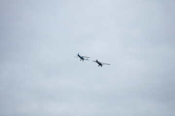 Two vintage-style airplanes are seen soaring in a pale sky.  A light blue and a dark brown/grey aircraft.