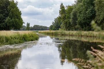 Tranquil canal, bordered by lush greenery and trees, reflects a cloudy sky.  Calm water, tranquil scene.