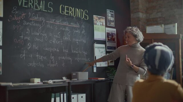 Experienced female teacher standing in sunlit college classroom, pointing at blackboard and explaining grammar rules to students in English class