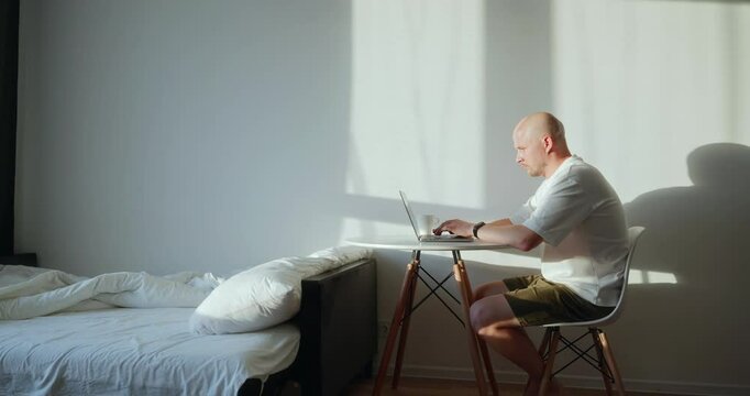A lonely bald introvert man works at home next to his bed at a table at dawn. A single workaholic man works at home next to an unmade bed. A workaholic's house is in disarray. High quality 4k footage