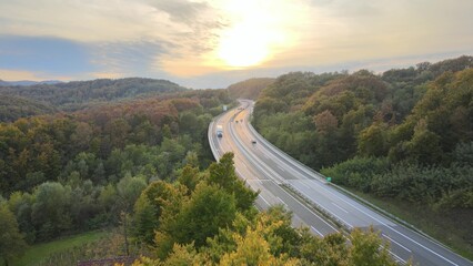 Aerial view of traffic highway through a green forest tree during colorful sunset autumn season. Green energy eco electric car no pollution