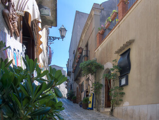 Charming cobblestone Erice street framed by lush greenery, historic buildings, and decorative...