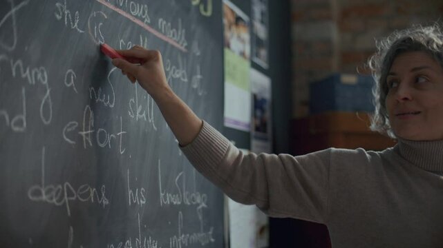 Middle-aged female teacher with curly grey hair pointing at text on blackboard and talking to students during English lesson in classroom