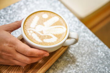 A woman holds a cup of latte art coffee at a cafe. A cup of latte coffee with art cream foam in a white ceramic cup on a wooden tray.