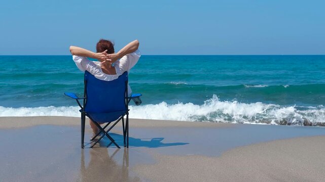 Enjoy your vacation by the ocean. Woman is enjoying a relaxing vacation day at the beach, sitting on a chair and stretching her arms while looking at the ocean