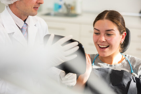 In dentists office, patient sits in dental chair with mirror in hands, see reflection, enjoys result of dental treatment. Male doctor dentist satisfied with result of prosthetics of girl patients