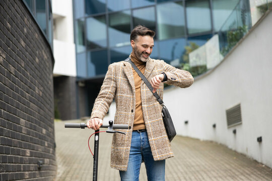 a stylish man in a checkered coat standing beside a scooter, checking his smartwatch with a modern urban background on a windy pathway.