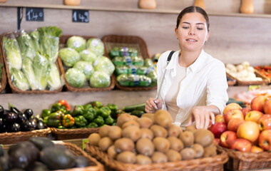 Delighted young woman purchaser choosing kiwi at the counter in large grocery store
