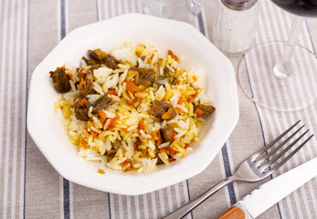 There is hexagonal plate with rice and chicken offal on striped tablecloth. Food is made from stewed chicken hearts, carrots, rice, spices.